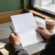 Hands holding a blank sheet of paper and a pen at a sunlit cafe table, a coffee cup beside them