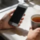 Hand holding a smartphone at a sunlit kitchen bench, the screen turned away, a cup of tea beside it, pausing before tapping