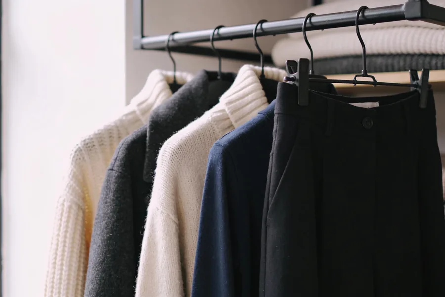 Small rail of winter clothes in a muted palette with a cream knit, a charcoal coat, a navy long-sleeve top and dark trousers, folded knits on a shelf behind