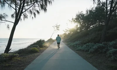 Runner seen from behind on a wide empty coastal path in Australia on a crisp winter morning, wearing light layers, with low coastal scrub and pale ocean beside the path