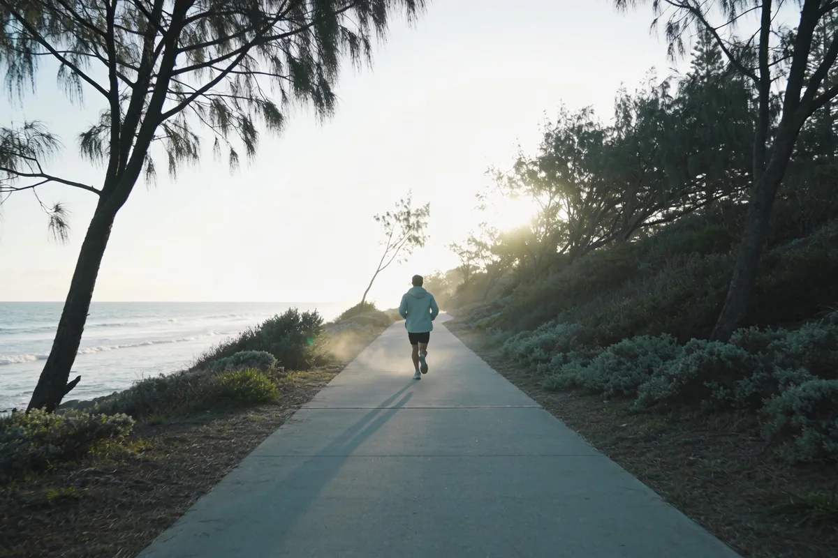 Runner seen from behind on a wide empty coastal path in Australia on a crisp winter morning, wearing light layers, with low coastal scrub and pale ocean beside the path
