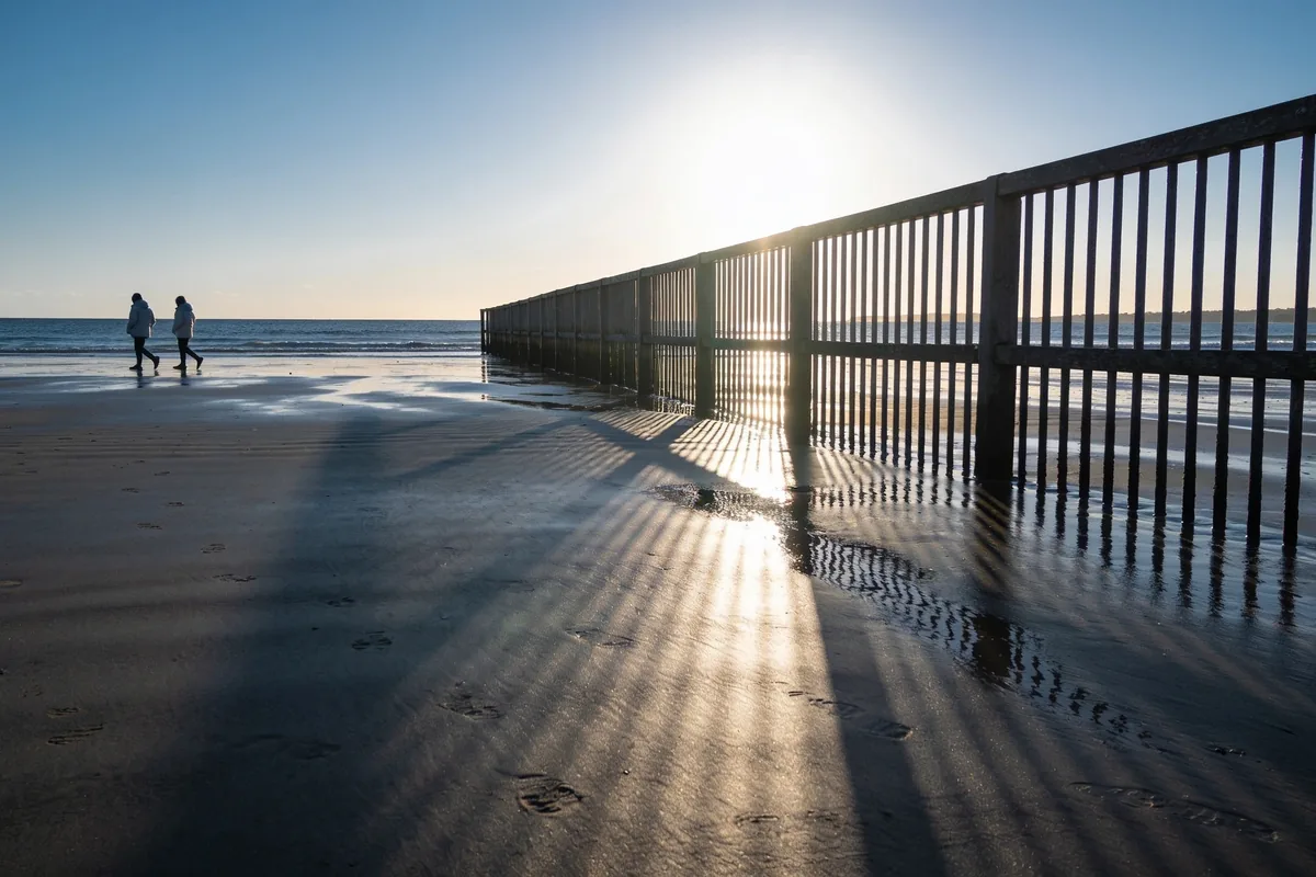 Empty Australian beach on a crisp winter morning with a low bright sun over the water and long shadows across damp sand, a couple walking in the distance in light jackets