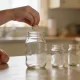 Child's hands placing a coin into a plain glass jar on a kitchen table in soft morning light, two other small glass jars beside it