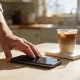 A hand resting on a smartphone lying face-down on a sunlit kitchen table beside a cold coffee and a notebook