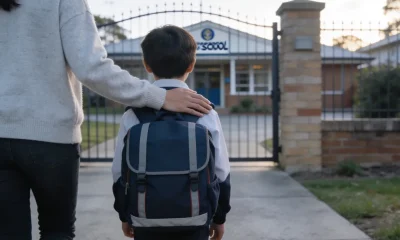 A parent and a primary-school-age child walking towards an Australian school gate, seen from behind