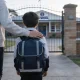 A parent and a primary-school-age child walking towards an Australian school gate, seen from behind
