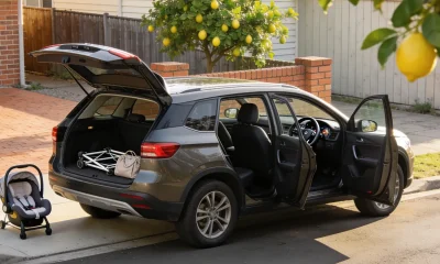 A clean mid-size family SUV parked at the kerb of a quiet Australian street on a bright morning with its tailgate and doors open, a child capsule standing on the footpath beside it and a folded pram frame and soft bag in the open boot, with a lemon tree, brick fence and weatherboard house behind
