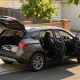 A clean mid-size family SUV parked at the kerb of a quiet Australian street on a bright morning with its tailgate and doors open, a child capsule standing on the footpath beside it and a folded pram frame and soft bag in the open boot, with a lemon tree, brick fence and weatherboard house behind
