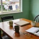A dining table in an ordinary Australian home set up for a meeting with three mismatched mugs, a plain closed folder, an open notebook with blank pages and a pen, and reading glasses, with chairs pulled up at angles and washing on a line in a suburban backyard beyond the window