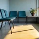 A bright empty waiting area in an ordinary Australian public building seen from one of the seats, with teal chairs against a bare pale wall, a low table with a single potted plant and hard mid-morning light across a vinyl floor