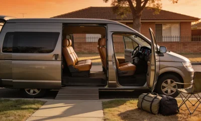 A plain silver-grey family van parked at the kerb of an outer-suburban street in late-afternoon light with its sliding and front doors open onto a cabin of tan seats, two soft travel bags and a folded camp chair on the grass verge beside it, with a brick-veneer house and street tree behind