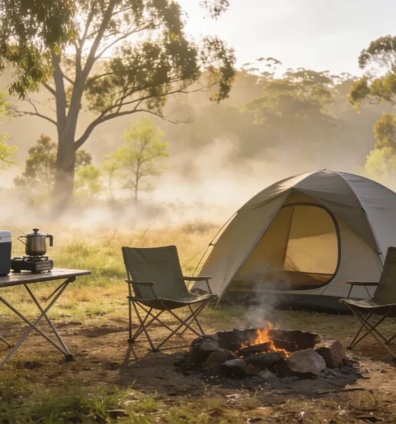 Simple dome tent at a small Australian spring campsite in early morning light, with two camp chairs by a fire ring, an esky and a camp stove on a folding table, eucalyptus trees behind.