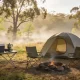 Simple dome tent at a small Australian spring campsite in early morning light, with two camp chairs by a fire ring, an esky and a camp stove on a folding table, eucalyptus trees behind.