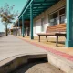 A quiet regional Victorian main street corner on a bright still morning seen from footpath level, with a low kerb and a gently sloped ramp, a timber bench seat beside a blank shopfront under a deep teal veranda and a clear path running away past a street tree