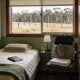 A quiet bedroom in an Australian holiday cottage seen from the doorway, with a made single bed and folded blanket, a plain closed book on the quilt, a soft open overnight bag and folded towels on a chair beside a lamp and a glass of water, and dry grass, a fence line and eucalyptus beyond the window