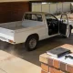 A white ute parked nose in under the open carport of an ordinary Australian suburban home in clear morning light, its driver's door standing open on the right hand side so the steering wheel, instrument dials and ignition key show inside on the right of the cabin, with a folder, a set of keys and a blank page on the low brick step beside a hose reel and a broom, and a colourbond fence and dry garden beyond