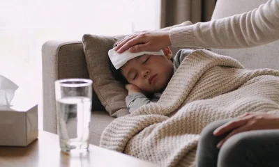 Parent placing a cool cloth on a young child's forehead while the child rests under a knitted blanket on a lounge, with a glass of water and tissues on the coffee table nearby.
