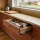 An ordinary Australian kitchen in mid morning light with a base cabinet drawer pulled open and sitting crooked, its shallow interior holding cutlery, a wooden spoon, a folded tea towel and a plain tin, with a kettle and a wooden board on the bench, a few crumbs scattered near a garden window and timber look flooring below