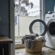 An Australian laundry on a weekday morning seen from the doorway, with the round door of a front loading washing machine standing open onto an empty drum, a woven basket of folded grey fleece and towels on the floor, a plain glass jar of washing powder with its lid on top of the machine, a wooden peg basket on a side bench and a clothesline against a paling fence and gum tree beyond the window