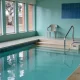 Calm water filling most of a rehabilitation pool room in Australia in soft daylight, with a wide shallow entry step and stainless steel handrail, a plain plastic step box at the water's edge, a folded towel and a water bottle beside a plastic chair on the dry tiled surround, and a quiet suburban street of brick fence and gum tree through the windows