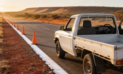 A long straight Northern Territory highway shoulder at sunrise, with plain orange traffic cones receding along the white edge line and a white ute parked on the shoulder seen from behind and to its left, its front left door standing open so the steering wheel, instrument dials and driver's seat show plainly across the cabin on the right hand side, with red gravel, scrub and a distant ridge under a low sun