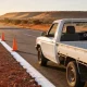 A long straight Northern Territory highway shoulder at sunrise, with plain orange traffic cones receding along the white edge line and a white ute parked on the shoulder seen from behind and to its left, its front left door standing open so the steering wheel, instrument dials and driver's seat show plainly across the cabin on the right hand side, with red gravel, scrub and a distant ridge under a low sun
