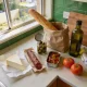 A pale kitchen benchtop in an ordinary Australian home on a bright weekend morning, freshly unpacked shopping on the surface: a wedge of pale cheese on greaseproof paper, a cured sausage on paper, an open jar of olives with its lid off, a shallow tin of roasted peppers, a paper bag with a long bread stick, a bottle of olive oil, a folded tea towel, two ripe tomatoes and a wooden board pushed aside, with a garden and paling fence through the window