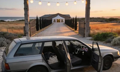 A silver grey station wagon parked at the gravel entrance of a rural Australian wedding venue at dusk, its nose pointing to the right of frame so its right hand driver's side faces the camera, the front and rear doors standing open on that near side so the steering wheel, steering column and instrument dials show plainly on the right of the cabin with the passenger seat beyond, a folded jacket and soft bag on the back seat, an open timber gateway with a string of lit festoon globes above a crushed rock driveway that runs past a cypress windbreak to a weatherboard hall and a white marquee under a soft evening sky