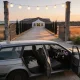 A silver grey station wagon parked at the gravel entrance of a rural Australian wedding venue at dusk, its nose pointing to the right of frame so its right hand driver's side faces the camera, the front and rear doors standing open on that near side so the steering wheel, steering column and instrument dials show plainly on the right of the cabin with the passenger seat beyond, a folded jacket and soft bag on the back seat, an open timber gateway with a string of lit festoon globes above a crushed rock driveway that runs past a cypress windbreak to a weatherboard hall and a white marquee under a soft evening sky