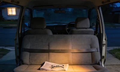 The back seat of a plain car at night seen from behind through the open rear of the cabin, a grey cloth bench seat with a seatbelt lying across it and a folded newspaper and pair of glasses resting on the cushion, the backs of the two empty front seats ahead and the steering wheel plainly on the right hand side of the frame beyond the passenger seat, with a quiet dark suburban street and one lit window seen through the windscreen and the interior light glowing warm over the seat