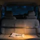 The back seat of a plain car at night seen from behind through the open rear of the cabin, a grey cloth bench seat with a seatbelt lying across it and a folded newspaper and pair of glasses resting on the cushion, the backs of the two empty front seats ahead and the steering wheel plainly on the right hand side of the frame beyond the passenger seat, with a quiet dark suburban street and one lit window seen through the windscreen and the interior light glowing warm over the seat