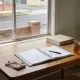 An open ring binder with blank pages and a pen on an ordinary Australian dining table in soft morning light, with a stack of plain envelopes, a mug of tea, folded reading glasses and a leafy houseplant at the edge of frame, a suburban street with a low brick fence and a street tree seen through the window behind