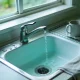 An ordinary Australian kitchen sink in the late morning, seen from just above bench height: a stainless steel single lever tap with clear water running in a steady stream into a plain white sink, a few drops already on the stainless steel beside the drain, a plain mug drying on the drainer and a tea towel folded over the edge of the laminate bench, with a soft green garden out of focus through the window above and ordinary weekday daylight across the whole scene