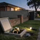 A suburban Australian front garden at dusk with the lid of a concrete water meter box lifted and leaning against a low brick fence, a brass water meter and tap fitting on copper pipe inside the open box, a torch lying lit on the wet grass beside a dark damp patch, and a brick veneer house, paling fence, wheelie bin and street tree behind under the first streetlight
