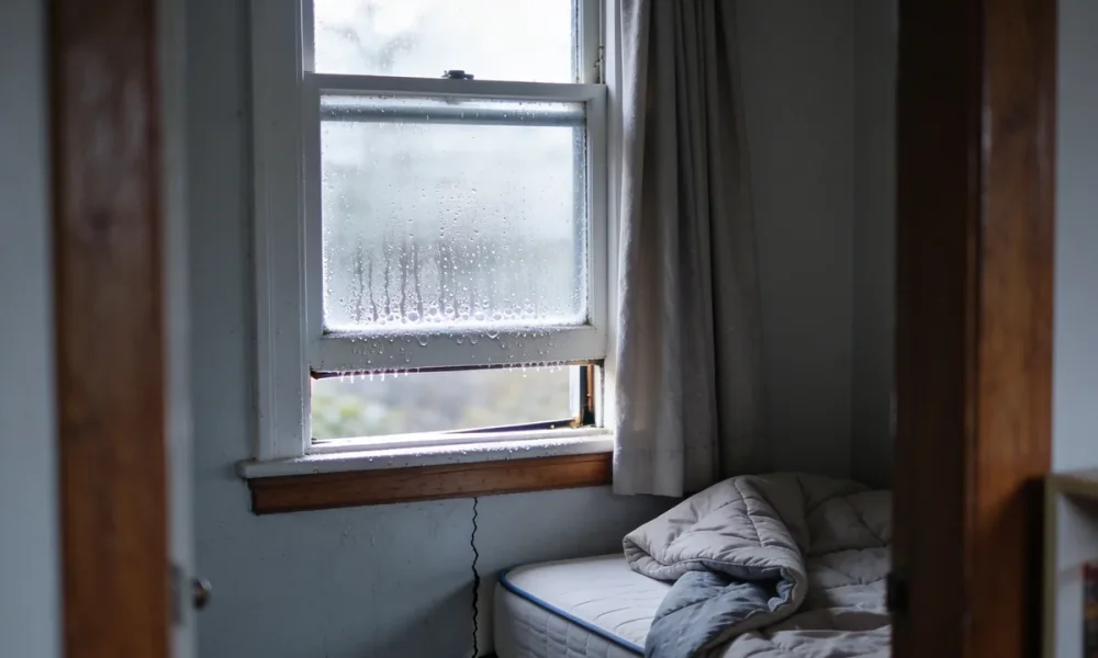 A corner of an ordinary Australian bedroom on a cold bright morning seen from the doorway, a sash window with its lower pane open a hand's width and condensation beaded along the glass and painted frame, a curtain drawn back to one side and a bed with the doona folded back standing a small gap from the cold wall, over timber floorboards with a low bookshelf at the edge of frame