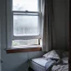 A corner of an ordinary Australian bedroom on a cold bright morning seen from the doorway, a sash window with its lower pane open a hand's width and condensation beaded along the glass and painted frame, a curtain drawn back to one side and a bed with the doona folded back standing a small gap from the cold wall, over timber floorboards with a low bookshelf at the edge of frame