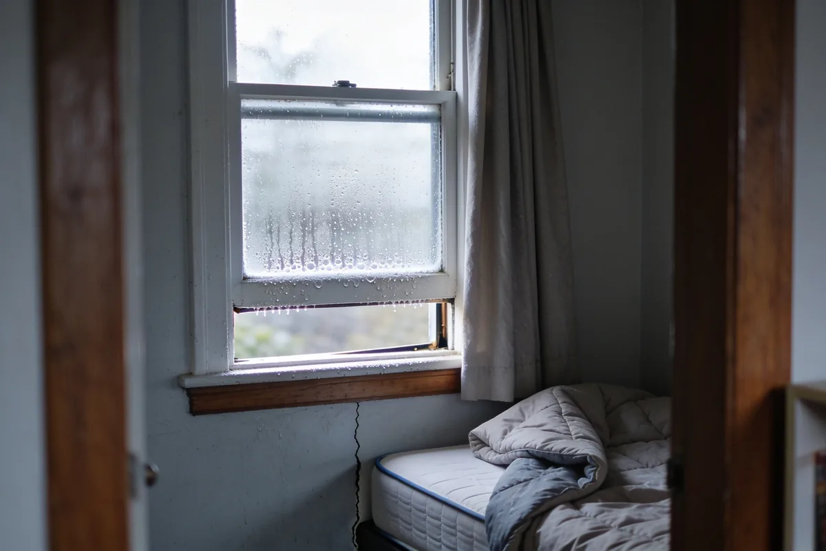 A corner of an ordinary Australian bedroom on a cold bright morning seen from the doorway, a sash window with its lower pane open a hand's width and condensation beaded along the glass and painted frame, a curtain drawn back to one side and a bed with the doona folded back standing a small gap from the cold wall, over timber floorboards with a low bookshelf at the edge of frame