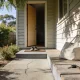 The front porch of an ordinary single storey Australian weatherboard home in the middle of a bright morning, seen from the front path: a plain timber front door standing open a hand's width onto a dim hallway with a pair of everyday shoes on the mat by the open door, a soft canvas bag standing on the porch boards beside a bunch of keys with a black fob lying on a plain ring, dappled shade from a leafy tree across the weathered boards and wall, a low native garden and a timber paling fence along the path, and a concrete path with a slight crack leading up to the step