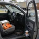 A plain unmarked grey work ute parked at the kerb of a quiet suburban Australian street at midday, seen from beside its open driver's door with the vehicle's nose pointing to the right of frame so its right hand driver's side faces the camera: the steering wheel, the driver's seat and the pedals of the near footwell plainly inside the open doorway with the instrument dials visible behind the wheel, and on that front seat a clipboard with faint ruled lines and empty boxes and nothing written on it lying across two plain unmarked cartons, a folded plain orange work vest with reflective stripes, a black pen, a neat coil of cable on the cabin floor and a takeaway coffee cup standing in the pocket of the open door, with a quiet street, a parked box truck and a low brick veneer house soft and out of focus beyond the windscreen