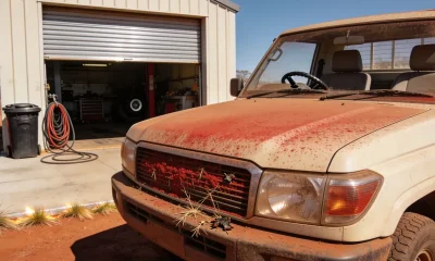 The front of a red dust covered Australian dual cab ute outside the part open roller door of a small outback workshop in the Pilbara on a hard bright day, its nose pointing to the left of frame so its left hand side faces the camera, the grille packed with red dust, dried grass seed and dead insects and the bonnet and windscreen filmed with red dust, the steering wheel sitting through the windscreen on the far side of the cabin at the vehicle's right hand driver's side ahead of the driver's seat, with a wheelie bin and coiled air hose at the workshop door, a concrete apron and red dirt and spinifex at the edge of frame