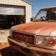 The front of a red dust covered Australian dual cab ute outside the part open roller door of a small outback workshop in the Pilbara on a hard bright day, its nose pointing to the left of frame so its left hand side faces the camera, the grille packed with red dust, dried grass seed and dead insects and the bonnet and windscreen filmed with red dust, the steering wheel sitting through the windscreen on the far side of the cabin at the vehicle's right hand driver's side ahead of the driver's seat, with a wheelie bin and coiled air hose at the workshop door, a concrete apron and red dirt and spinifex at the edge of frame