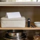An open kitchen cupboard in an ordinary Australian home in soft morning light holding a plain cream soft sided zip case with a folded blank leaflet standing on top of it, a small black torch, a pair of scissors and a white enamel bowl beside it and stacked stainless steel saucepans and a jar of dried goods on the shelf below