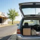 The open boot of a plain silver station wagon parked at the kerb of a quiet country town main street on a clear morning, seen from behind and slightly to one side so its clean unbranded nose points away from the camera: a single plain closed cardboard carton, a soft canvas bag and a folded striped blanket sitting in the open load space under the raised tailgate, and looking forward through the open rear of the cabin past the empty passenger seat, the steering wheel and the driver's seat sit plainly on the right hand side of the cabin at the right of frame, with a low row of plain single storey shopfronts with awnings and dark empty windows along the left, a leafy street tree, a wide almost empty road and a hint of golden paddocks at the end of the street