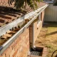 The eave line of a single storey Australian brick veneer house seen from the garden in late afternoon light, galvanised guttering holding shallow water with dry broad leaves and seed pods along the channel above a downpipe running down the brickwork into a grated pit, a tiled roof and an overhanging gum tree branch above, with dry lawn, a paling fence and a hose reel at the edge of frame