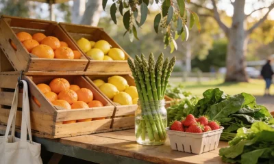 Spring fruit and vegetables piled on a wooden market stall at an Australian farmers market, with citrus crates, asparagus standing in water and a punnet of strawberries