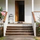 The front verandah of a single storey Queenslander style home on a bright morning, seen from the front path: wide timber steps up to a verandah with simple timber handrails and white posts, a plain timber front door standing open onto a dark hallway, two pairs of everyday shoes on the mat by the door, and a small plain wheeled walking frame standing against the wall beside the door with no markings on it, with a frangipani in white and pink flower on one side and a red flowering bottlebrush on the other along a low timber fence, and dry grass at the edge of the frame
