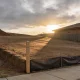 A cleared suburban building block on a slight slope in the late afternoon, seen from the footpath of the street it fronts: bare graded earth with a shallow batter falling away to the left, a low ridge of stockpiled soil along one boundary, a new plain timber survey peg standing at the corner with no markings on it, a temporary sediment fence of dark fabric running along the low edge on plain posts, and a single storey brick veneer house of similar vintage seen over the boundary fence to the right, with long shadows across the earth, dry grass at the margins and an overcast sky broken by low late sun