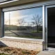 The outside of a suburban Australian brick veneer house on a cool bright winter morning, seen from the front garden at a slight angle: a large fixed window and an adjacent sliding glass door in a plain aluminium frame, both clean and closed, the window carrying a clear reflection of the street's bare winter trees, a power pole and open sky so nothing behind the glass is legible and the darker glass of the door showing only a dim undefined room beyond, with a low brick front wall, a native garden bed with dry mulch and a concrete path leading to the door, and cold early light with long shadows across the lawn and brickwork