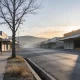 An empty country town main street early on a clear winter morning, seen from the footpath: a wide empty road with no vehicles at all, low rows of single storey shop buildings with plain awnings and dark empty windows down both sides, a large bare winter street tree standing at the kerb near the camera, dry golden grass in the gutter and along the kerb, and a low ridge of hills at the end of the street clearing out of the dawn haze under a soft warm sky, with long shadows lying across the road